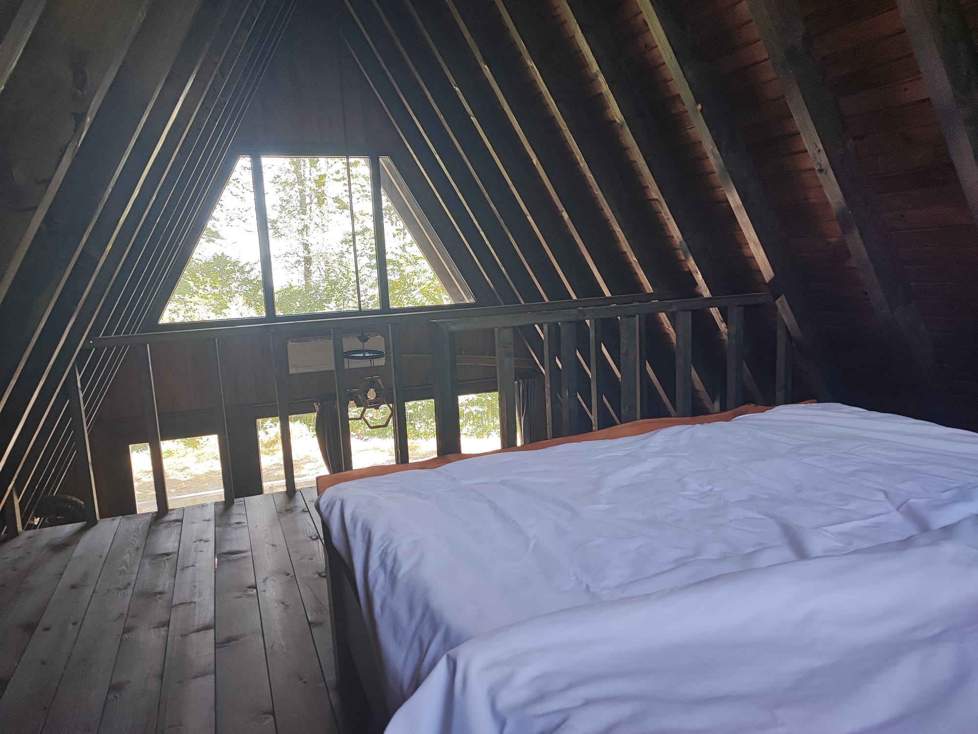 Loft bedroom with a white-linen bed on a timber-railed mezzanine directly beneath the triangular apex window, looking out to the forest.