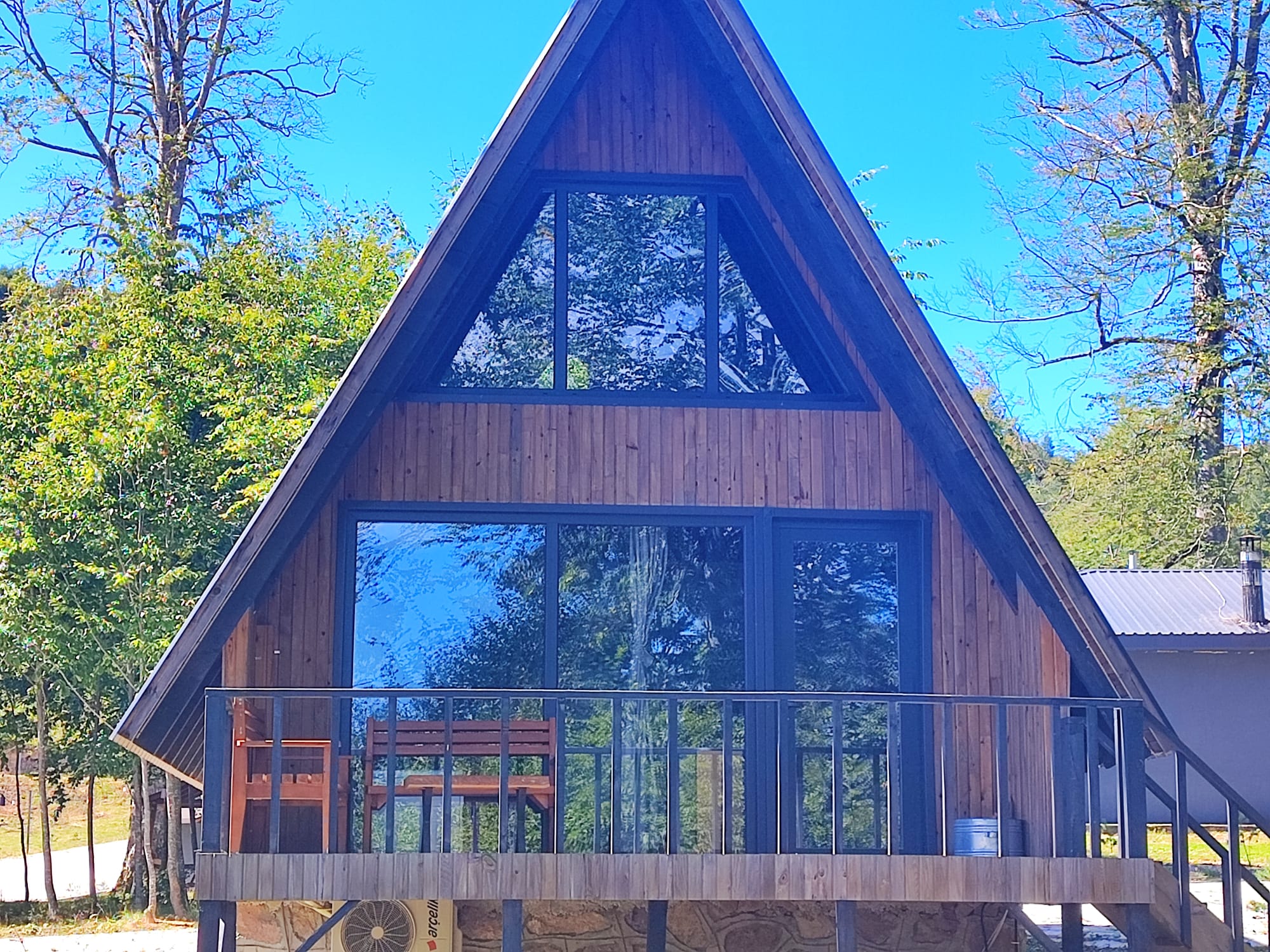 The A-frame bungalow seen head-on, its full triangular glass gable reflecting the surrounding forest above a timber deck.