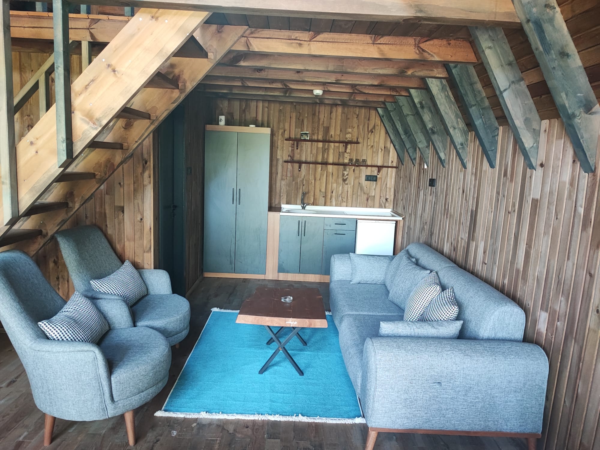 Living space with a grey sofa and armchairs around a teal rug and wood table, with a compact kitchenette tucked beneath the loft stair.