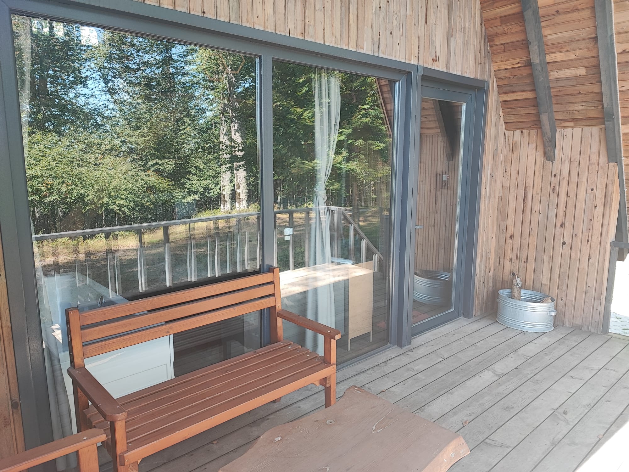 A furnished terrace with a wooden bench and grey decking, the sliding glass doors reflecting the surrounding forest.