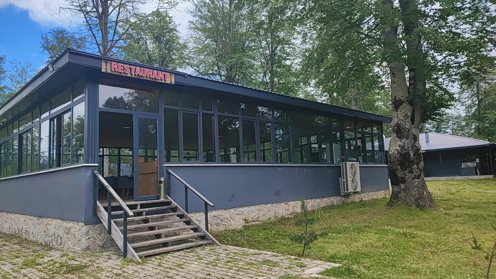 The dark-framed glass restaurant building seen from outside, its lit Restaurant sign above timber steps, set among tall trees.