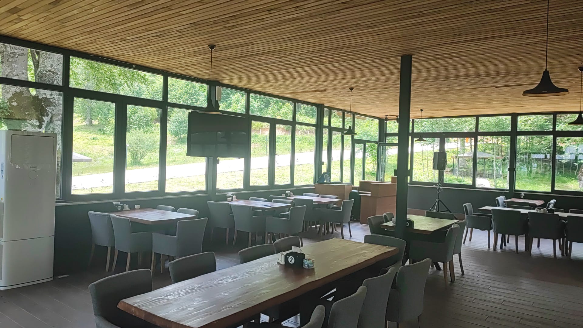 A second view of the glass dining room with long communal wood tables and grey chairs, lit by green daylight from the surrounding trees.